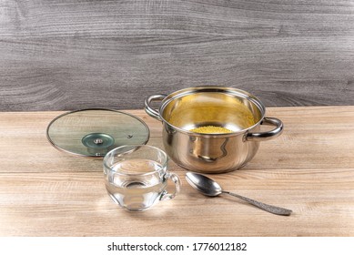 A Metal Pan With A Glass Lid Contains Millet Grits, Salt, Sugar And A Mug Of Water For Cooking Millet Porridge On A Wooden Table