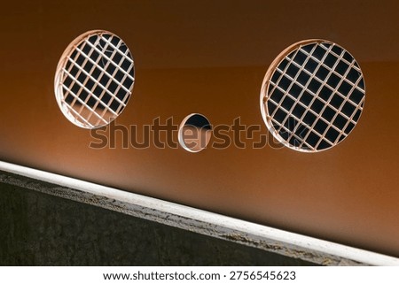 Metal panel featuring two circular ventilation grilles with grid patterns and a small round hole in the center, illuminated by soft lighting.