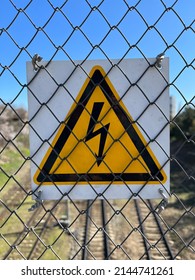 Metal plate representing high voltage warning sign as extra safety measures in industry. Closeup of high voltage warning sign mounted on steel wire mesh against blue sky and railways on the background