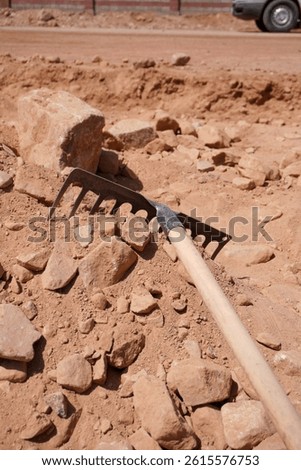 A metal rake rests on a rocky surface, surrounded by various sizes of stones and dirt. The background shows a blurred vehicle and a dusty road.