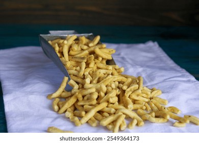 A metal scoop pours golden, crispy snack sticks onto a white cloth, highlighting their texture and color.
