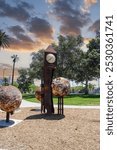 a metal sculpture of a grandfather clock and three large rusty balls in the park surrounded by lush green palm trees and grass and purple trees with powerful clouds at sunset at MacArthur Park	