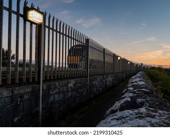 Metal Security Fence With Light And Passing Train. Dark And Moody Sunset Light. Travel Concept. Safety Matter.