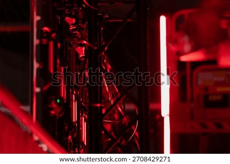 Metal truss structure with mounted technical equipment and visible cabling illuminated by intense red lighting, forming a complex arrangement within a dark industrial or stage-oriented workspace