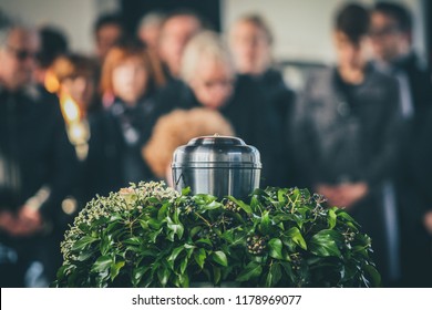 A metal urn with ashes of a dead person on a funeral, with people mourning in the background on a memorial service. Sad grieving moment at the end of a life. Last farewell to a person in an urn. - Shutterstock ID 1178969077