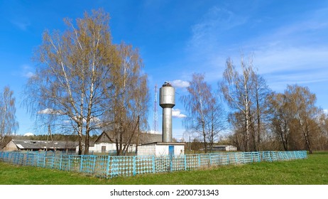 A Metal Water Tower With A Nest And Storks On Top Is Enclosed By A Metal Fence. Nearby Are Birch Trees Then There Are Farm Buildings A Mobile Operator Tower And A Forest It's Sunny And The Sky Is Blue