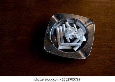 A metallic ashtray on a wooden surface contains several cigarette butts and a crumpled piece of paper, highlighting themes of smoking, waste, and addiction. - Powered by Shutterstock