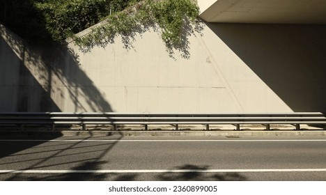 Metallic guardrail on sidewalk. Concrete wall of an underpass, with vegetation at top, on behind and two lane road in front. Background for copy space