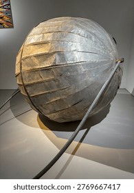 Metallic spherical object with segmented panels. It rests on a polished floor surface. Cable extends from its side toward the foreground. Soft lighting highlights its weathered texture and form
