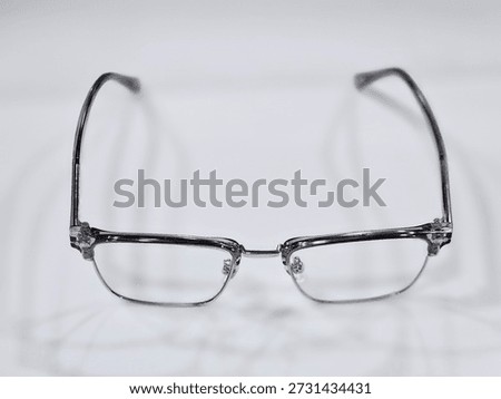 A metallic-framed pair of rectangular eyeglasses with nose pads lies on a white surface, casting a soft shadow that highlights its minimalist design and structure.