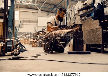 A metallurgy worker kneeling at workshop and processing metal with grinder.