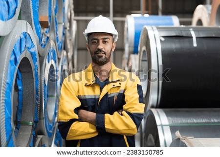 Metalwork manufacturing, warehouse of raw materials. Portrait of male factory worker standing with crossed arms near rolls of metal sheet in aluminum material warehouse