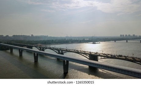 Metro Bridge and Communal Bridge. Two parallel crossings span the wide Ob River in Novosibirsk, Russia, with the covered metro structure in the foreground and the older arched Communal Bridge carrying road traffic behind it. The scene is captured from an elevated aerial perspective, showing the steel and concrete constructions stretching across the broad waterway under a bright, hazy summer sky. The river surface reflects sunlight in shimmering patterns, while the distant cityscape and industrial skyline fade into the atmospheric haze. The metro crossing, recognized as the longest covered metro span in the world at 2145 meters, features a distinctive enclosed gallery design that protects trains from Siberian weather extremes. The Communal crossing displays classic arched metal construction with multiple spans and heavy vehicle traffic. Together these structures form a vital transport corridor connecting the two banks of the Ob in this major Siberian city. The overall atmosphere is one of functional industrial grandeur set against the natural expanse of the waterway, with warm daylight and soft clouds overhead.