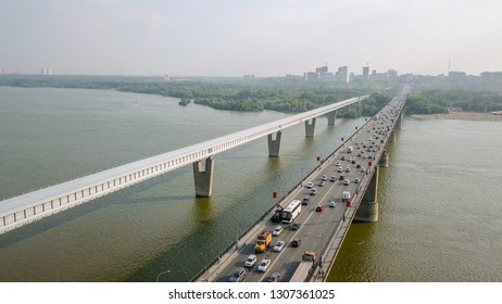 Metro Bridge and Communal Bridge. Two parallel spans cross the wide Ob River in Novosibirsk, Russia, with the enclosed white metro structure running alongside the multi-lane road crossing carrying dense automobile traffic. The metro span, the longest covered metro crossing in the world at 2,145 meters, features a continuous steel beam structure with reinforced concrete pillars and glazed side galleries. The adjacent Communal crossing, built in 1955, serves as a vital road artery with vehicles flowing in both directions. The scene is set on a hazy summer day with flat, diffused light over the broad river, forested banks, and the distant city skyline. Shot from an elevated aerial perspective, the composition emphasizes the engineering scale and the contrast between the clean white transit structure and the busy grey road crossing. Travel, transportation, landmark, cityscape.