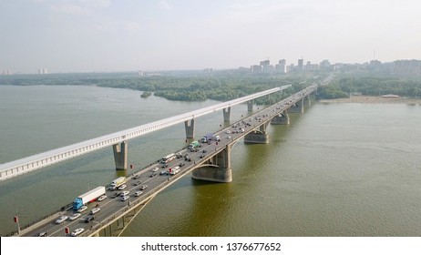 Metro Bridge and Communal Bridge. Two parallel structures span the wide Ob River in Novosibirsk, Russia, with the covered railway crossing running alongside the multi-lane roadway carrying heavy vehicle traffic. The enclosed steel beam structure, sheathed in a white gallery, holds the record as the longest covered metro crossing in the world at 2,145 meters total length, with its main span stretching 896 meters across the river channel. The adjacent Communal Bridge carries a steady flow of cars and trucks across reinforced concrete piers. Both structures cross the seventh-longest river in the world, connecting the left and right banks of Siberia's largest city. The scene is captured from an elevated aerial perspective on a bright summer day, with green riverbanks, distant city buildings, and a pale sky forming the backdrop. The white gallery contrasts sharply with the darker road deck and the greenish-brown waters below, creating a striking engineering landmark in an urban riverside setting.