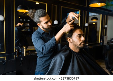 Mexican barber using scissors to give a new haircut and hairstyle to a handsome caucasian client at the barber shop - Powered by Shutterstock