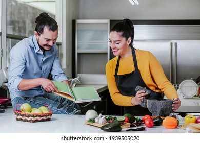 Mexican Couple Reading Book With Recipes While Cooking Mexican Food In Kitchen At Home In Mexico City