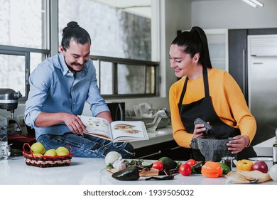 Mexican Couple Reading Book With Recipes While Cooking Mexican Food In Kitchen At Home In Mexico City