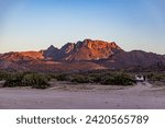 Mexican desert landscape with juniper trees and scrubland, imposing mountain rock formation illuminated by sun in background, car parked on sand, sunset in desert of Baja California Sur, Mexico