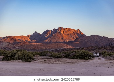 Mexican desert landscape with juniper trees and scrubland, imposing mountain rock formation illuminated by sun in background, car parked on sand, sunset in desert of Baja California Sur, Mexico - Powered by Shutterstock