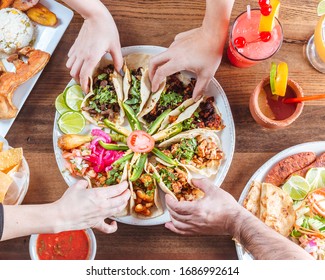 Mexican Family Taco Plater On Dark Wood Suraface With Four People Reaching Into Frame To Grab Tacos With Traditional Mexican Drinks And Salsa And Beans And Fajitas