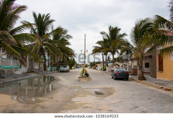 Mexican fishing village street during the rainy\
season with cars parked along the sides and bag of garbage in the\
middle of the street by\
pole