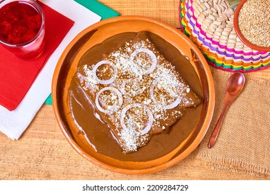 Mexican Food (mole Enchiladas) Served With Onion Rings, Aged Cheese And Sesame Seeds In A Clay Dish On A Wooden Table With Colorful Folkloric Decorations. Top View