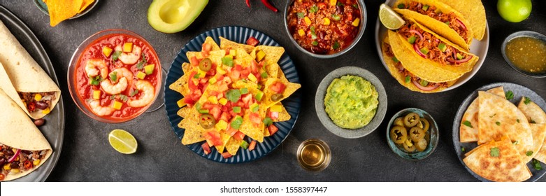 Mexican Food Overhead Panoramic Shot, Many Dishes Of The Cuisine Of Mexico, Flatlay On A Dark Background. Nachos, Guacamole, Shrimp Cocktail, Tacos, Chili Con Carne