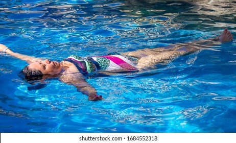 Mexican Mature Woman Floats Relaxing In A Swimming Pool With Crystal Clear Water On A Blue Background, Black Hair, Swimsuit With White, Pink, Green And Black Colors, Sunny Day Of Relaxation