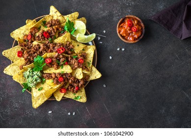 Mexican nacho corn tortilla chips with cheese, meat, guacamole and red hot spicy salsa. Nachos with ground beef on dark rustic background, copy space.