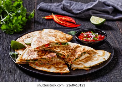mexican quesadilla filled with chicken, green onion, cheese, chilli pepper, coriander leaves served with tomato salsa and lime on black plate on dark wood table