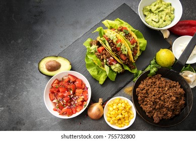 mexican tacos and ingredients like fried ground beef, tomato salsa, guacamole, corn and spices on a dark slate plate with copy space, top view from above, selected focus