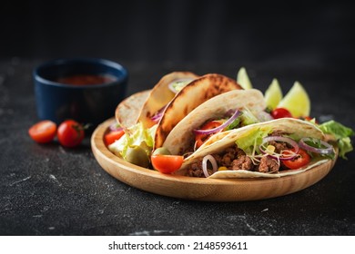 Mexican Tacos In Yellow Corn Tortillas Filled With Carnitas, Onion, Tomatoes And Salad Served In Wooden Plate On The Black Concrete Background