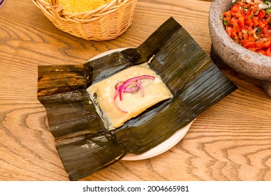 Mexican tamale of cochinita pibil stewed inside corn cob leaf with volcanic stone molcajete filled with pico de gallo