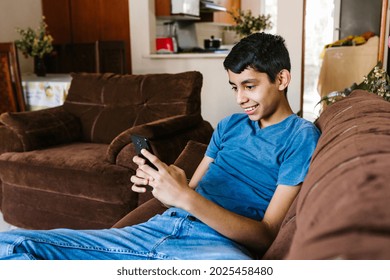 Mexican Teen Boy Having Fun With Smartphone Sitting On Sofa At Home In Latin America