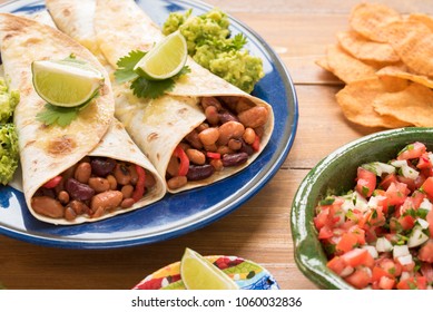 Mexican three bean, flour tortilla wraps with guacamole, salsa, lime wedges, coriander and tortilla chips on a wooden background