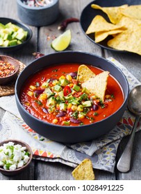 Mexican Tomato, Bean, Bell Pepper Soup In Black Bowl With Nachos Chips. Wooden Background.