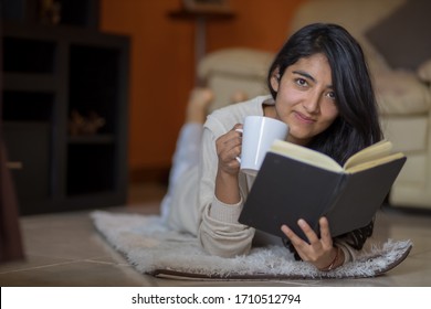 Mexican Woman  Lying Down Reading A Book At Home Drinking Coffee Due To Coronavirus Quarantine