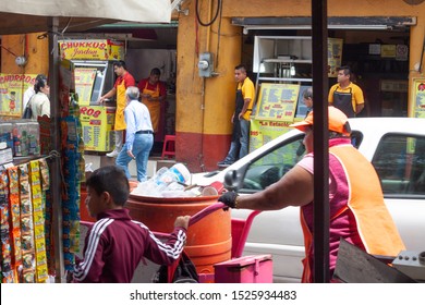 Mexico City, Mexico, September 01, 2018: View Of The Street From Inside A Store, With A Boy And A Girl Picking Up Trash And Food Stores On The Other Side. Travel Concept