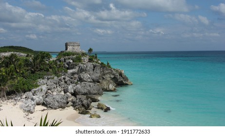 Mexico, Yucatan, Tulum Ruins - Ancient Maya Temple Of The God Of Winds On The Beach, With Clear Turquoise Water And Cloudy Sky Rocks And Vegetation, Seen From Afar 