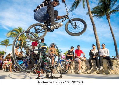 MIAMI - DECEMBER 27, 2017: A Young Daredevil Cyclist Performs A Stunt Jump Over Another Bicycle In Front Of Spectators On The Beachfront Boardwalk Promenade At Lummus Park In South Beach.