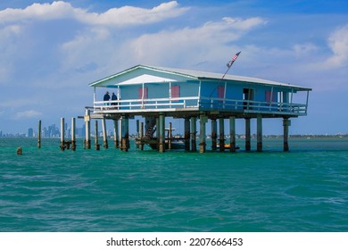 MIAMI, FL - APRIL 2, 2022: Historic Collection Of Homes On Stilts - Known As Stiltsville - In Biscane National Park With Cape Florida Lighthouse And Miami Skyline In The Distance
