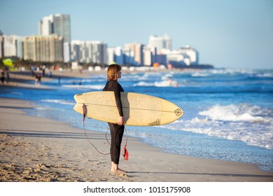 MIAMI - JANUARY 4, 2018: A Young Female Surfer In A Wet Suit Stands On The Shore Of South Beach With Her Surfboard Looking Out At The Winter Swells.