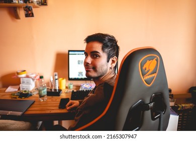 Miami, USA - 2022: Cougar Gamer Chair (in Gray And Orange Colors) And Young And Handsome Caucasian Boy With Earring In Front Of A Computer, Big Desk And Peach Wall