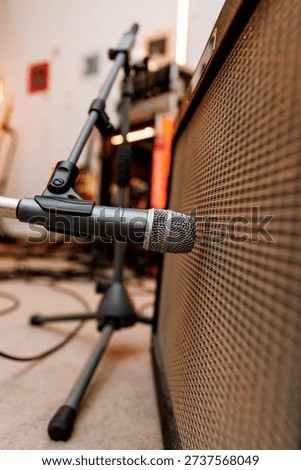 A microphone mounted on a stand faces a guitar amplifier inside a rehearsal room with cables in background.