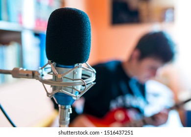 Microphone On Focus, Playing Young Guitarist On Background In A Small Music Studio, Shallow Depth Of Field.