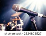 Microphone on stage – live showbiz music setting featuring close up of vocal mic for singing. Drum kit and stage lighting in background. Performance venue such as concert, festival, night club or bar.