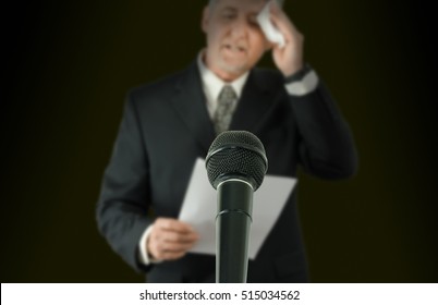 Microphone In Sharp Focus With A Nervous Sweaty Public Speaker Or Politician Blurred In The Background Preparing To Make A Speech As He Wipes His Brow