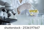 Microscope is near a scientist, wearing a lab coat and white protective gloves, and holding beaker with a yellow oily liquid in laboratory setting, close up. Medicine and science concept