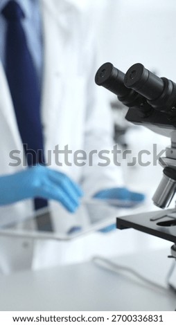 Microscope is on a table, ready for innovative medical research in a sterile laboratory environment. In the background, a scientist analyzes the data obtained. Concept of medicine and science